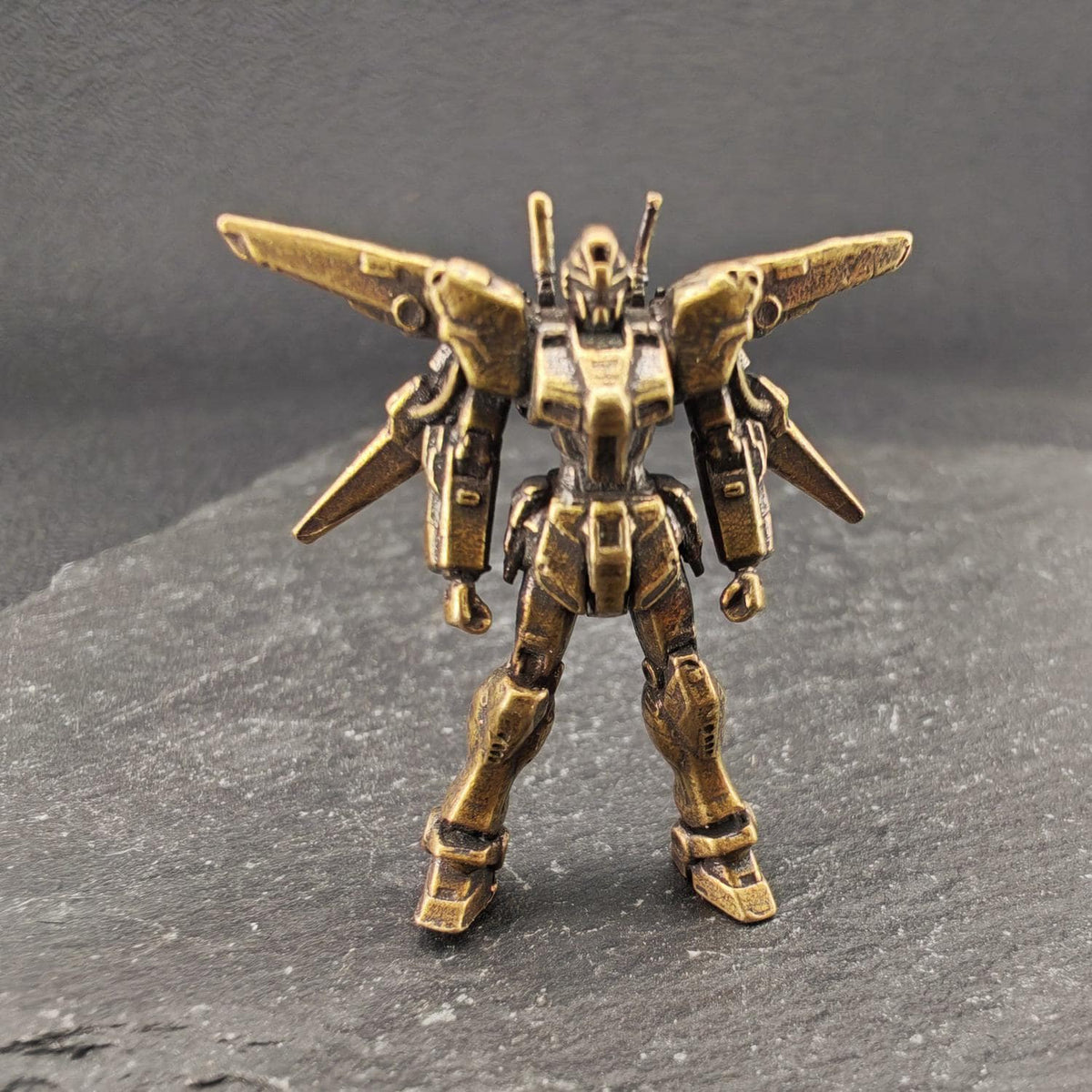 A small metallic gold robot action figure or miniature model standing on a dark textured surface against a dark gray background.