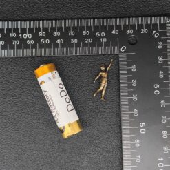 A small metallic gold figurine of a child standing on a black textured surface, shown next to a yellow and white Dodo brand cylindrical item and metal rulers for scale.